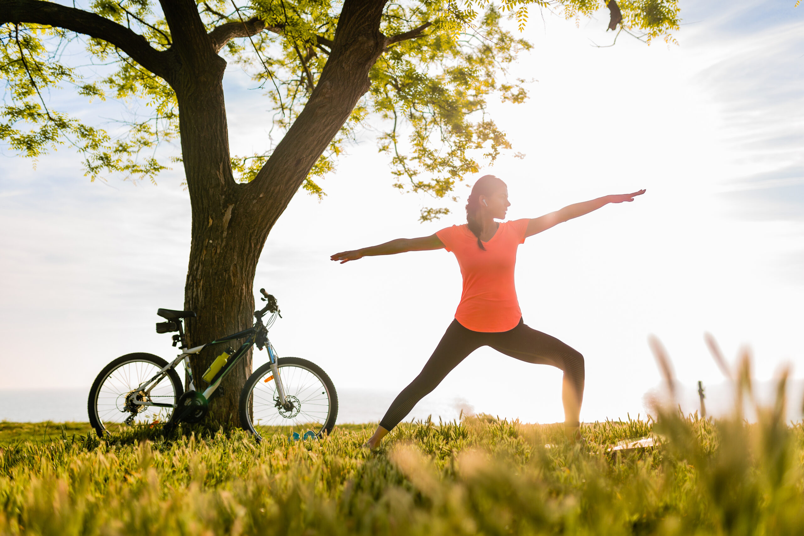 slim beautiful woman silhouette doing sports in morning in park doing yoga on mat in colorful fitness outfit in nature, smiling happy healthy lifestyle, calm meditation, bicycle on background