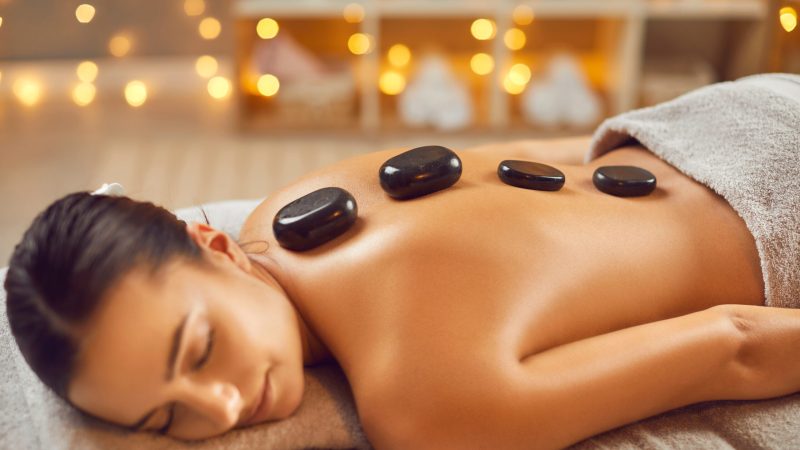 Beautiful serene young woman having spa hot stone massage. Relaxed brunette girl lying on couch with black basalt stones along spine. Beauty treatment therapy, body care concept