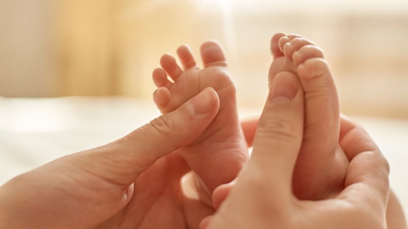 Mum making baby massage, mother massaging infant bare foot, preventive massage for newborn, mommy stroking the baby's feet with both hands on light background.