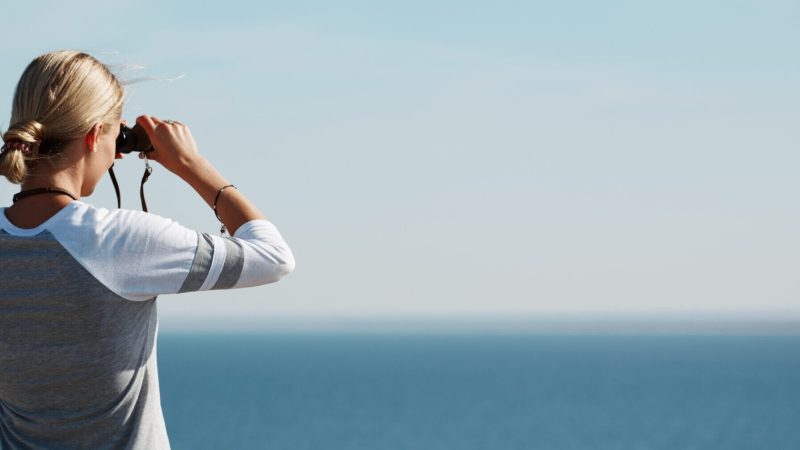 Woman tourist looking through binoculars at distant sea, enjoying landscape